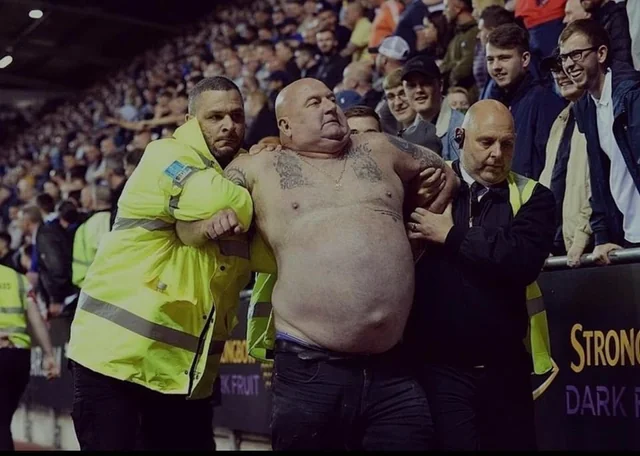 Funny photo of shirtless man being escorted by security at a crowded event — funny people pics caught at the perfect moment