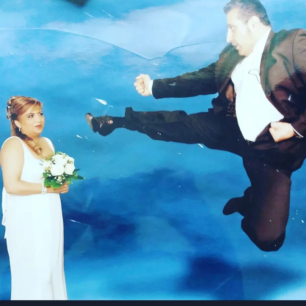 Groom in a tuxedo mid-air delivering a karate kick toward the bride holding flowers, both posing in front of a blue backdrop