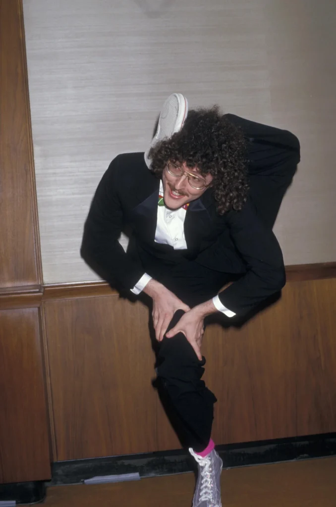 Weird Al Yankovic in a tuxedo, balancing with one leg bent up over his shoulder in an exaggerated pose