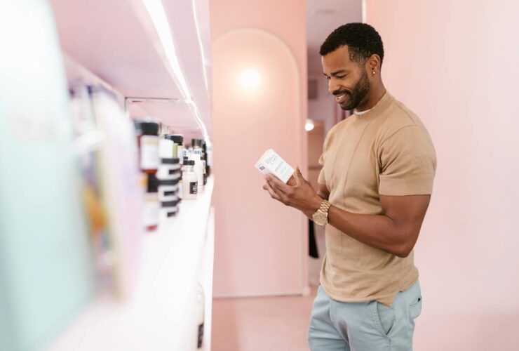 A man in a brown crew-neck T-shirt reading a wellness product label in a pastel-colored store. Represents informed and health-conscious shopping decisions.