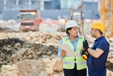 A man and a woman wearing hard hats discussing plans at a construction site.
