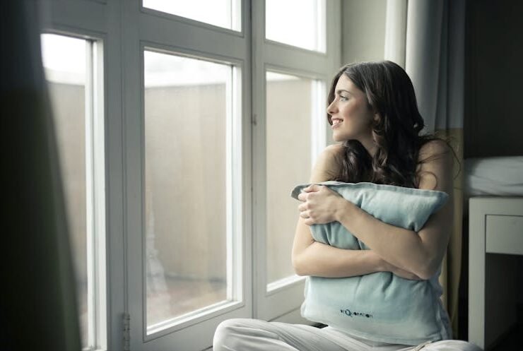Woman hugging a gray pillow near a window, preparing for seasonal home changes.